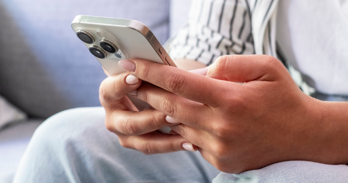 Nahaufnahme einer sitzenden Person in Jeans, die ein silberfarbenes Smartphone mit beiden Händen bedient.