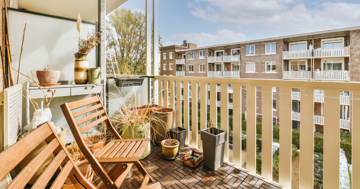 Kleiner, sonniger Balkon mit Holzboden, zwei Holzstühlen, verschiedenen Pflanzgefäßen und dekorativen Vasen; im Hintergrund sind die Balkone eines mehrstöckigen Wohnhauses zu sehen.