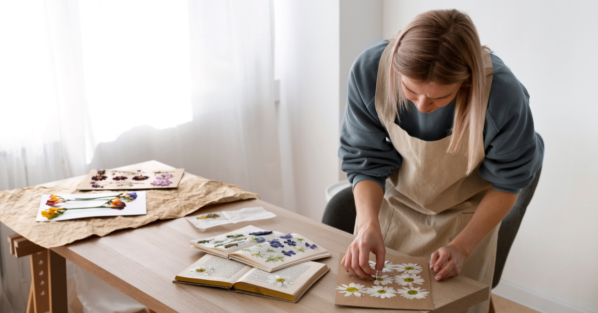 Eine Frau in Schürze arbeitet an einem Tisch und arrangiert gepresste Blumen auf einer Pappe. Auf dem Tisch liegen weitere gepresste Blüten und geöffnete Bücher mit Pflanzenmustern.