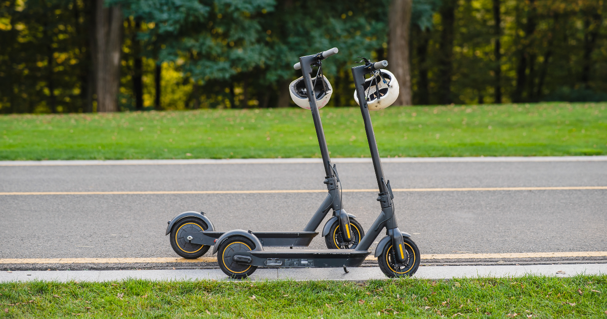 Zwei geparkte E-Scooter mit angehängten Fahrradhelmen stehen am Straßenrand vor einer Grünfläche mit Bäumen im Hintergrund.