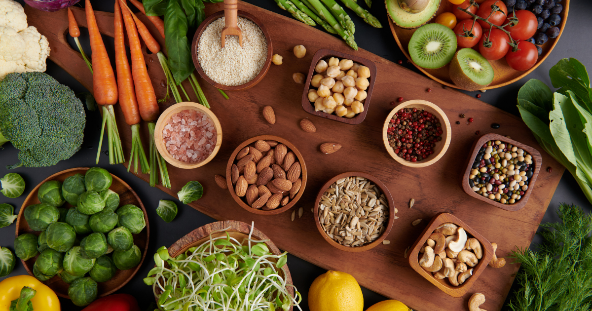 Verschiedene frische Lebensmittel wie buntes Gemüse, Obst, Nüsse, Samen und Hülsenfrüchte sind dekorativ auf einem großen Holzbrett arrangiert und bilden eine farbenreiche, gesunde Auswahl pflanzlicher Zutaten.