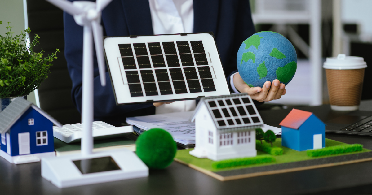 Person in Businesskleidung sitzt an einem Schreibtisch und hält ein Modell eines Solarpanels sowie eine kleine Erdkugel. Auf dem Tisch stehen Miniaturhäuser mit Solardächern, ein Modell-Windrad, Pflanzen und weitere Symbole für erneuerbare Energien.