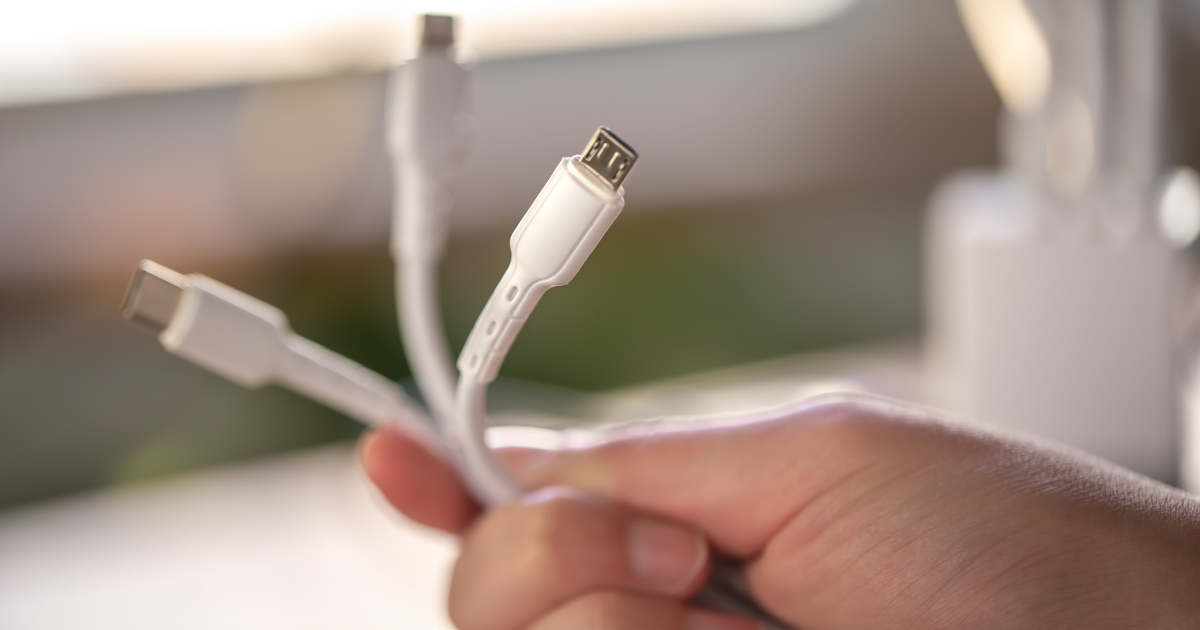 Hand hält mehrere weiße Ladekabel mit verschiedenen Steckertypen, darunter USB-C und Micro-USB, vor unscharfem Hintergrund.