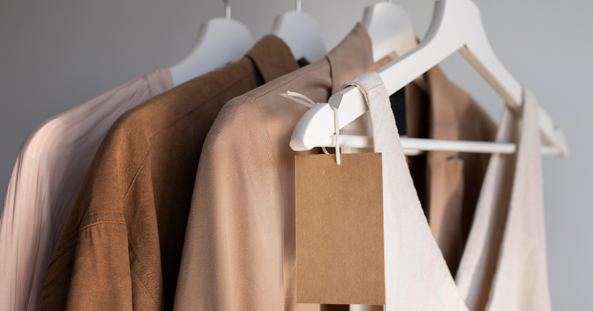 Mehrere Kleidungsstücke in neutralen Beige- und Brauntönen hängen ordentlich auf weißen Kleiderbügeln, eines davon mit einem schlichten braunen Papieretikett versehen.