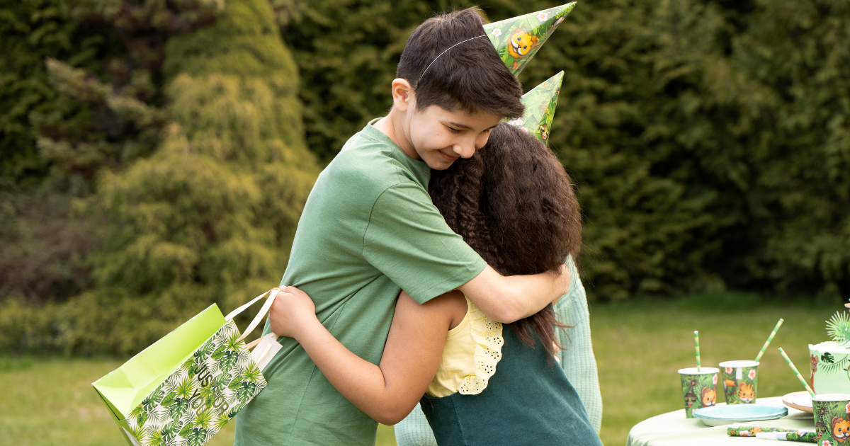 Zwei Kinder mit Partyhütchen umarmen sich im Garten; eines hält eine Geschenktüte, im Hintergrund steht ein gedeckter Geburtstagstisch mit grüner Deko.