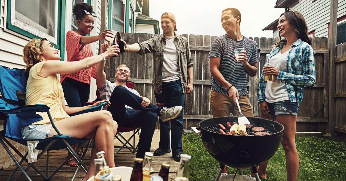 Eine Gruppe junger Erwachsener sitzt und steht fröhlich im Garten, während sie gemeinsam grillen. Einige stoßen mit Getränken an, andere unterhalten sich, und auf dem Grill liegen verschiedene Speisen. Die Szene wirkt entspannt und sommerlich.