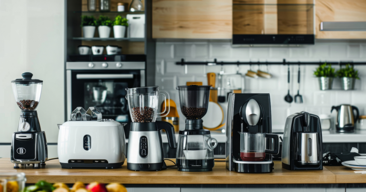 Mehrere Küchenmaschinen – darunter Mixer, Toaster, Kaffeemühlen, eine Kaffeemaschine und ein Wasserkocher – stehen ordentlich aufgereiht auf einer Arbeitsplatte in einer modernen Küche. Im Hintergrund sind Küchenschränke, ein Backofen und Regale mit Pflanzen und Utensilien zu sehen.