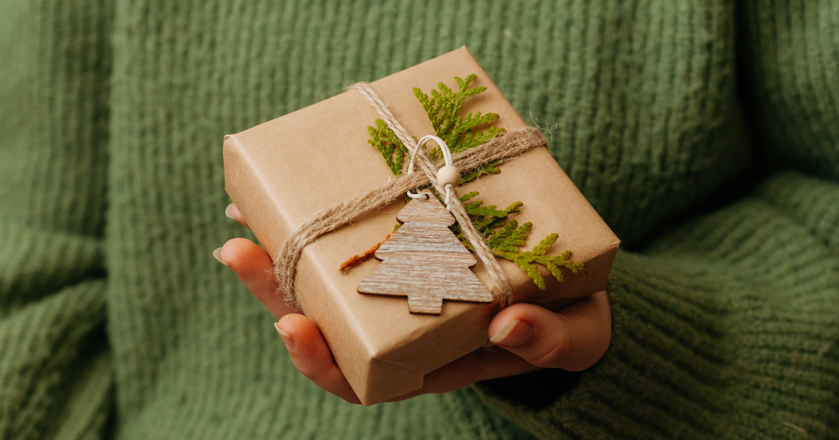 Hände halten ein naturbelassen verpacktes Geschenk mit braunem Papier, Juteschnur, grünem Zweig und einem kleinen Holzanhänger in Tannenbaumform.