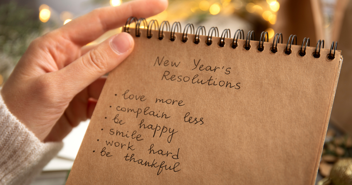 Hand hält ein Notizbuch mit der Überschrift „New Year's Resolutions“ und einer Liste guter Vorsätze, umgeben von warmen Lichtern im Hintergrund.