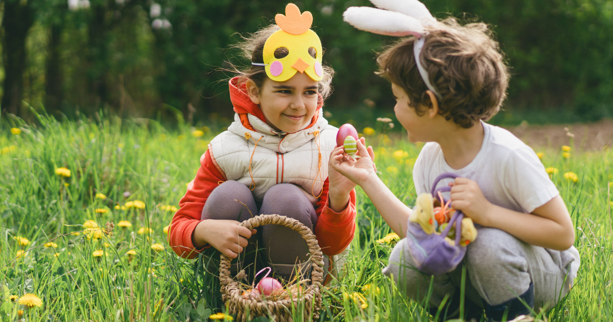 Zwei Kinder mit Osterkostümen suchen im grünen Gras nach Ostereiern. Ein Kind trägt eine gelbe Kükenmaske, das andere Hasenohren, und sie halten bunte Eier in den Händen.