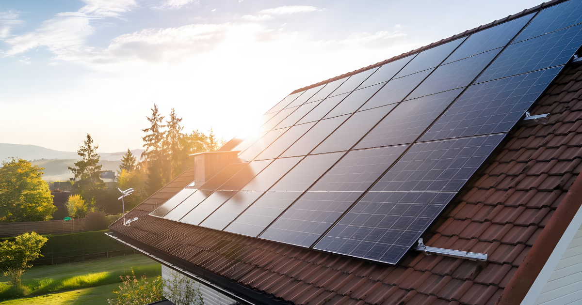Solaranlage auf dem Dach eines Hauses bei Sonnenaufgang, umgeben von grüner Landschaft und Bäumen.