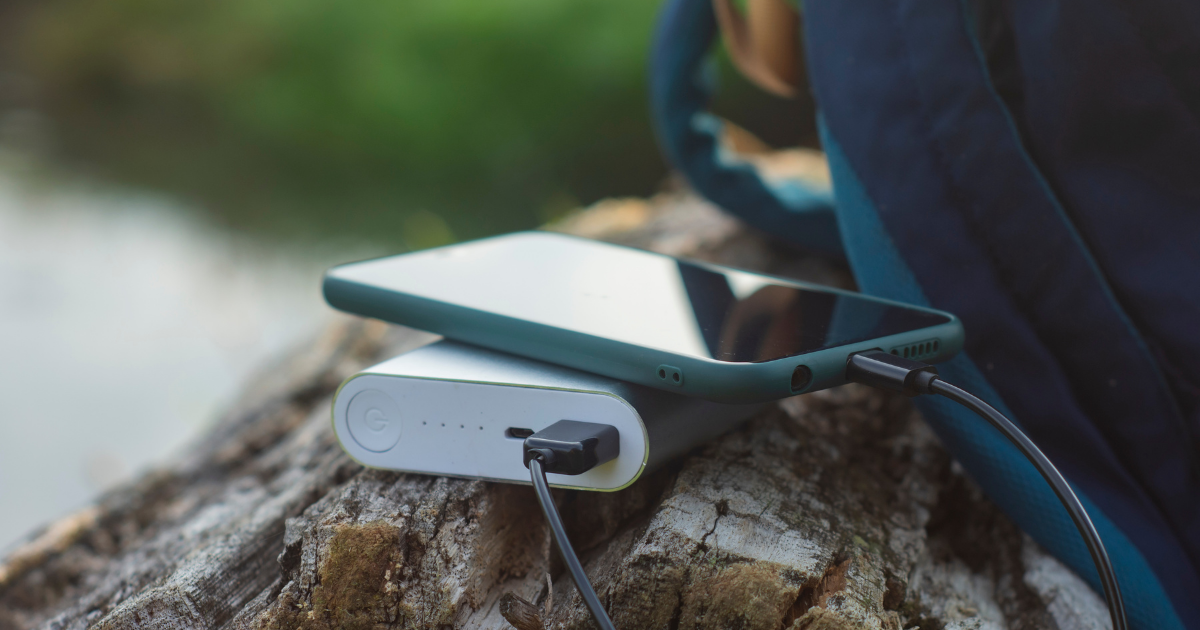 Ein Smartphone liegt auf einer Powerbank, die über ein Kabel angeschlossen ist. Beides befindet sich auf einem Baumstumpf im Freien, neben einem Rucksack.
