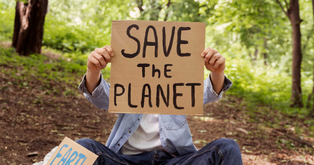 Eine sitzende Person hält ein Pappschild mit der Aufschrift „SAVE THE PLANET“ vor ihrem Gesicht, umgeben von Bäumen in einem Wald; neben ihr liegt ein weiteres Schild mit Umweltbotschaft.