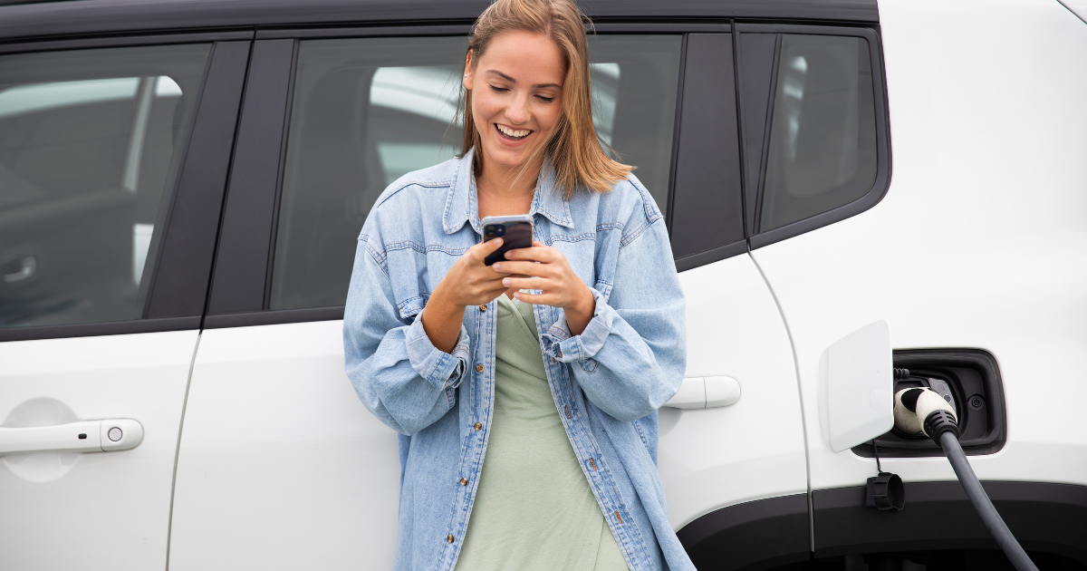 Eine lächelnde Person lehnt an einem weißen Elektroauto, das gerade geladen wird, und schaut auf ihr Smartphone.