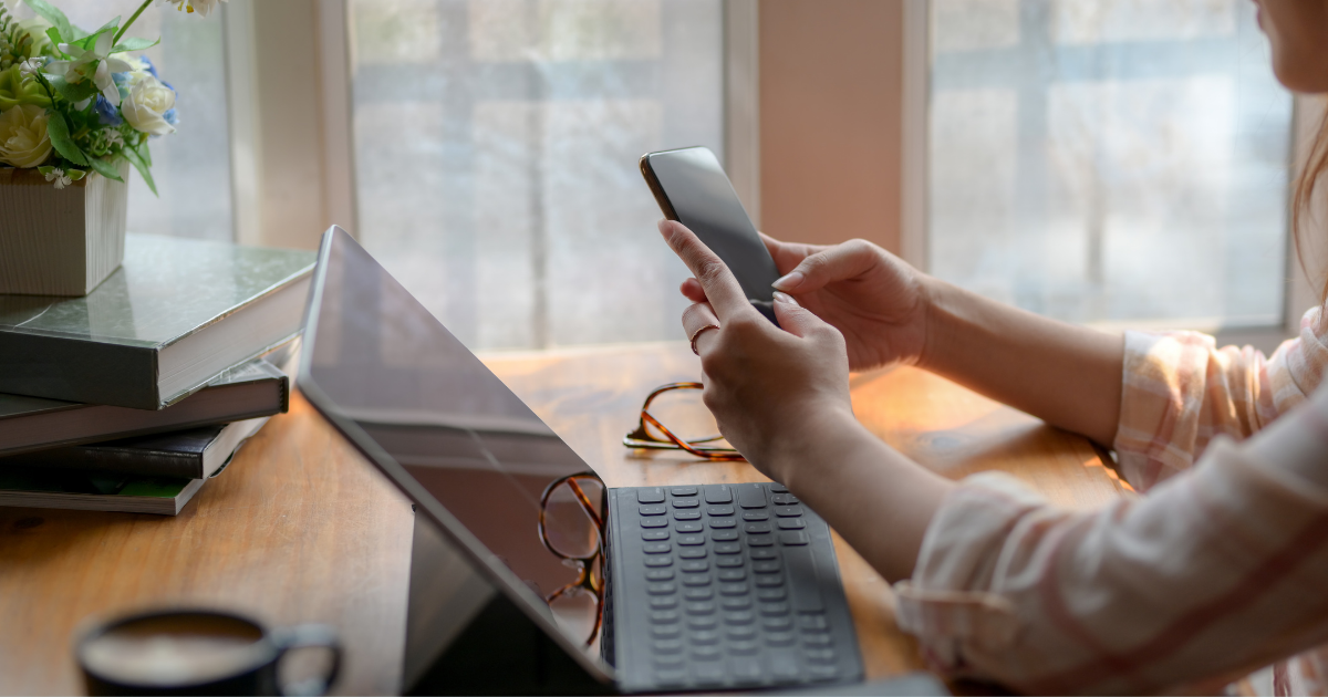 Person sitzt am Schreibtisch vor einem aufgeklappten Laptop und hält ein Smartphone in den Händen; auf dem Tisch liegen Bücher, eine Brille und eine Tasse Kaffee, im Hintergrund große Fenster mit Tageslicht.