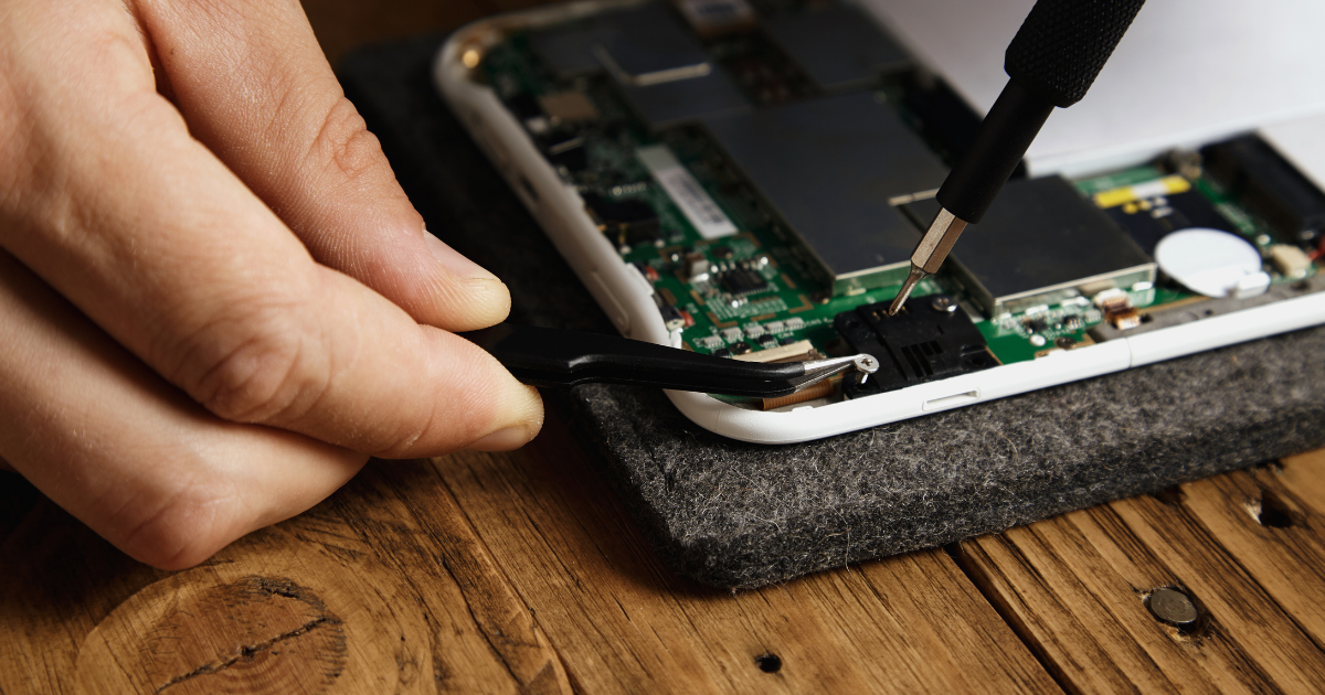 Nahaufnahme von Händen, die mit Werkzeugen ein geöffnetes Smartphone reparieren; interne Elektronik und Platinen sind sichtbar.