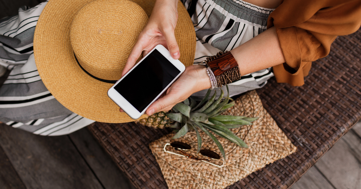 Person sitzt entspannt draußen, hält ein Smartphone in den Händen und hat einen großen Strohhut auf dem Schoß; daneben liegen eine geflochtene Tasche, eine Sonnenbrille und eine Ananas.