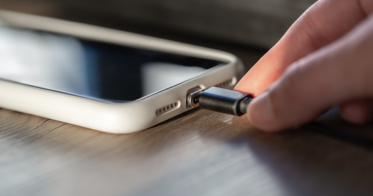 Nahaufnahme einer Hand, die ein Ladekabel in den Anschluss eines Smartphones mit weißer Hülle steckt, liegend auf einer Holzoberfläche.