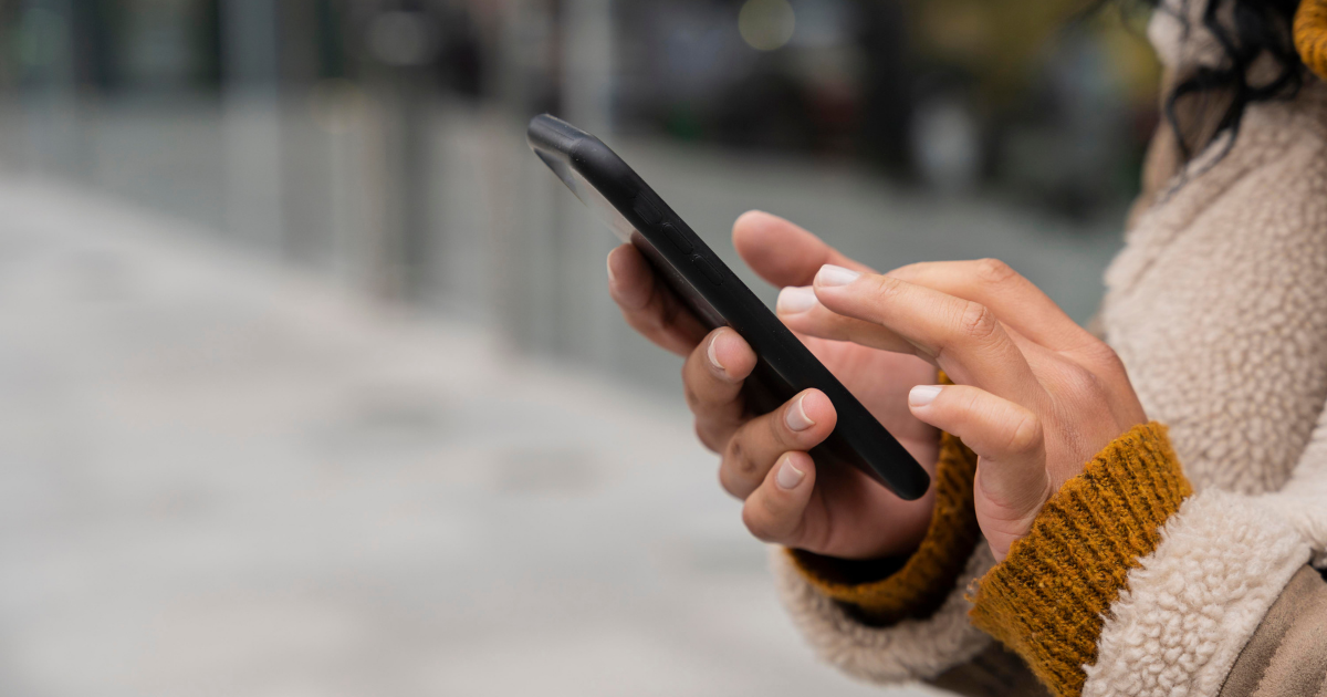 Nahaufnahme von Händen einer Person, die ein Smartphone hält und den Bildschirm berührt; unscharfer Hintergrund im Freien.