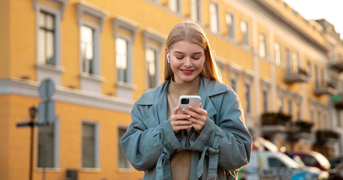Junge Frau mit Kopfhörern schaut lächelnd auf ihr Smartphone, während sie auf einer sonnigen Straße vor einem gelben Gebäude steht.