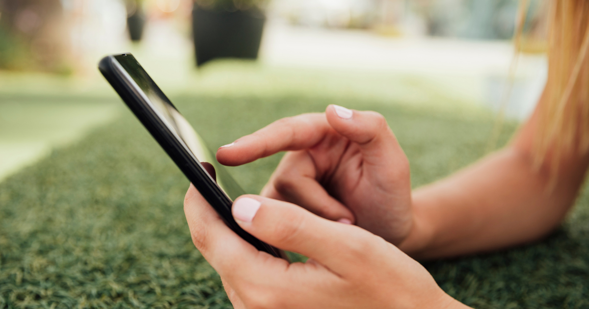 Nahaufnahme von Händen, die ein Smartphone halten und den Bildschirm berühren; im Hintergrund unscharfe Grünfläche im Freien.
