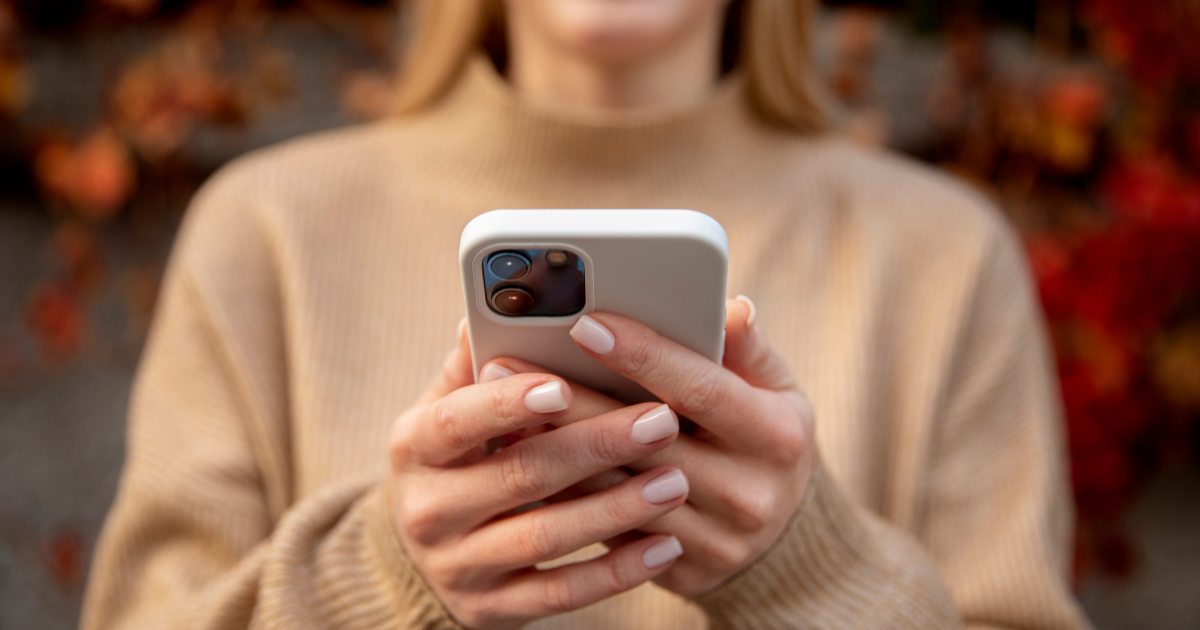 Eine lächelnde Person in einem beigefarbenen Pullover hält ein Smartphone mit beiden Händen.