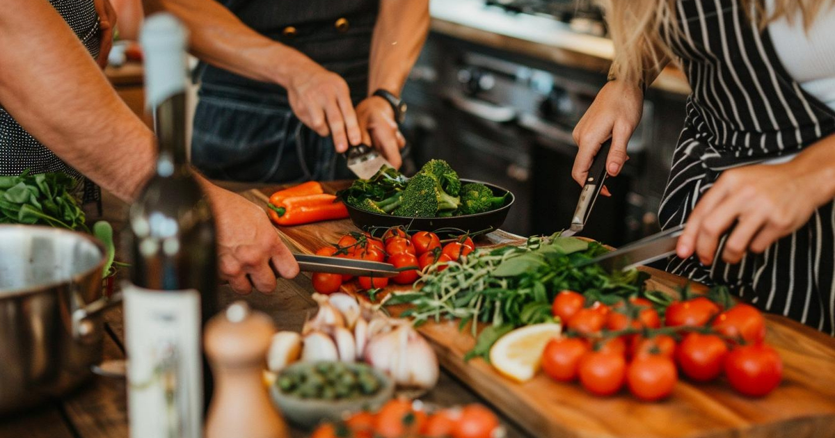 Mehrere Personen schneiden gemeinsam frisches Gemüse wie Tomaten, Kräuter, Paprika und Brokkoli in einer Küche; Kochutensilien und Zutaten liegen auf dem Holztisch bereit.