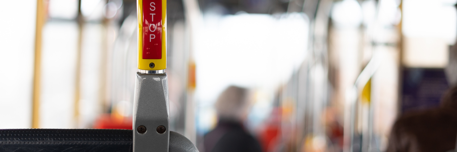 Nahaufnahme einer „Stop“-Taste im Innenraum eines Busses; im Hintergrund sind unscharf mehrere sitzende Fahrgäste und Haltestangen zu erkennen.