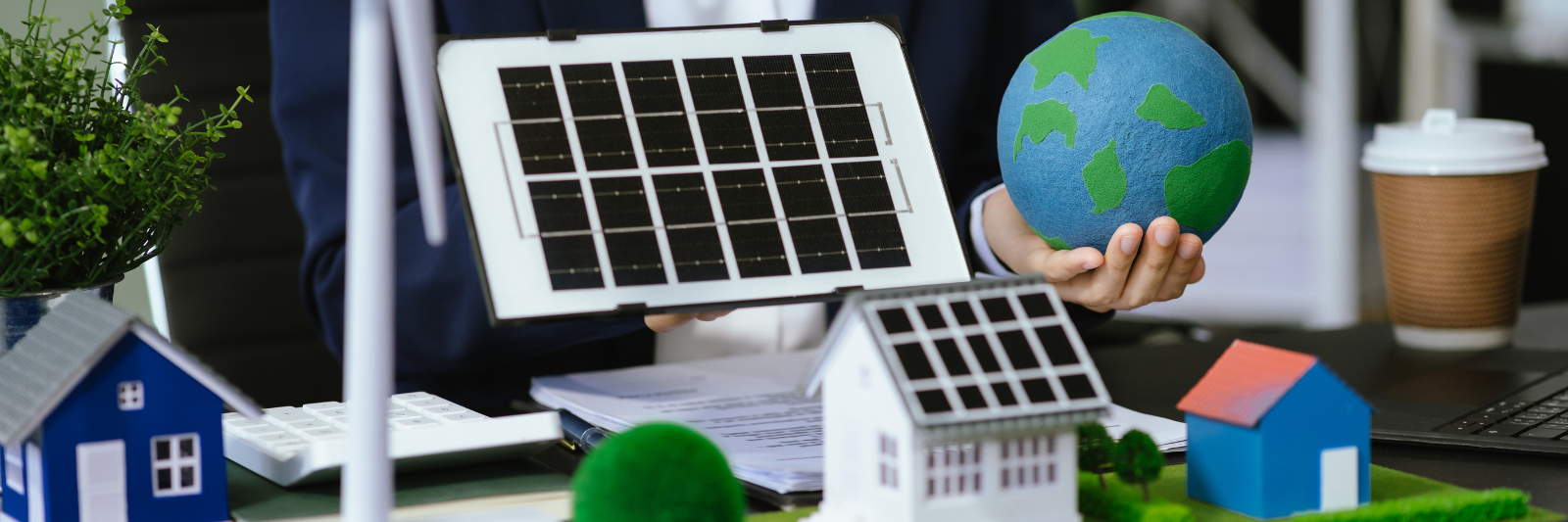 Person in Businesskleidung sitzt an einem Schreibtisch und hält ein Modell eines Solarpanels sowie eine kleine Erdkugel. Auf dem Tisch stehen Miniaturhäuser mit Solardächern, ein Modell-Windrad, Pflanzen und weitere Symbole für erneuerbare Energien.
