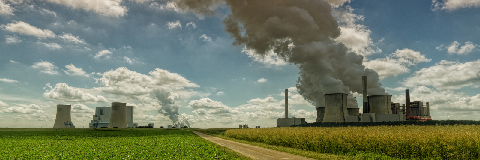 Großes Kraftwerk mit mehreren Kühltürmen, aus denen dichte Rauch- und Dampfwolken aufsteigen; davor Felder und ein Weg unter blauem Himmel mit Wolken.