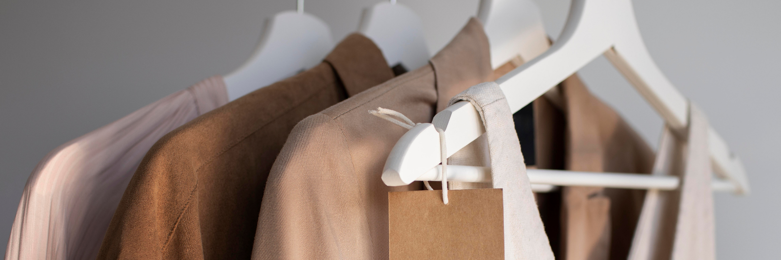 Mehrere Kleidungsstücke in neutralen Beige- und Brauntönen hängen ordentlich auf weißen Kleiderbügeln, eines davon mit einem schlichten braunen Papieretikett versehen.
