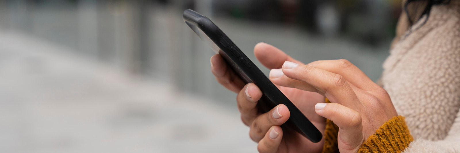 Nahaufnahme von Händen einer Person, die ein Smartphone hält und den Bildschirm berührt; unscharfer Hintergrund im Freien.