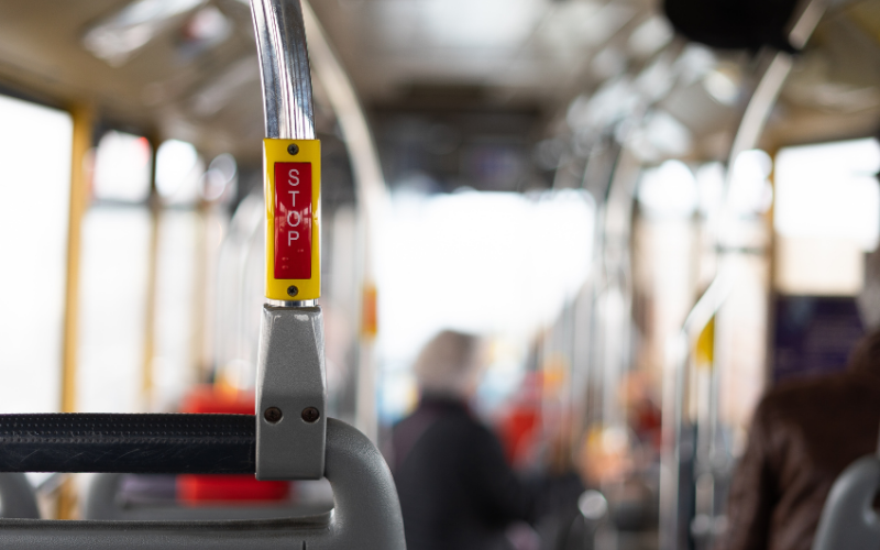 Nahaufnahme einer „Stop“-Taste im Innenraum eines Busses; im Hintergrund sind unscharf mehrere sitzende Fahrgäste und Haltestangen zu erkennen.
