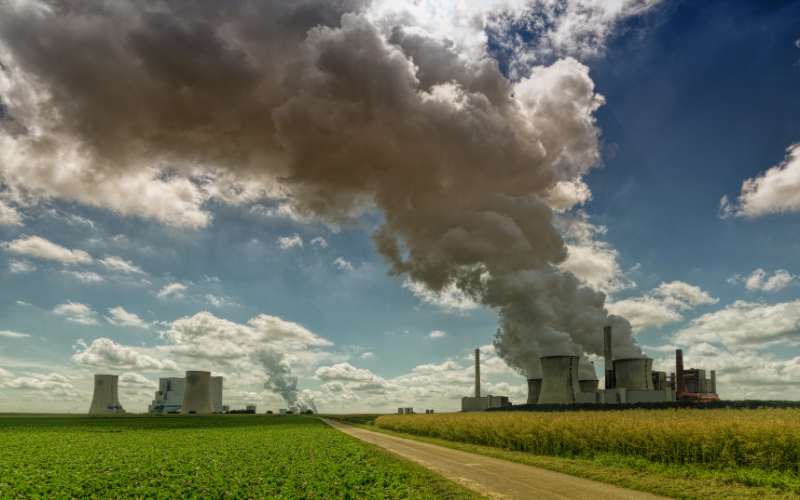 Großes Kraftwerk mit mehreren Kühltürmen, aus denen dichte Rauch- und Dampfwolken aufsteigen; davor Felder und ein Weg unter blauem Himmel mit Wolken.
