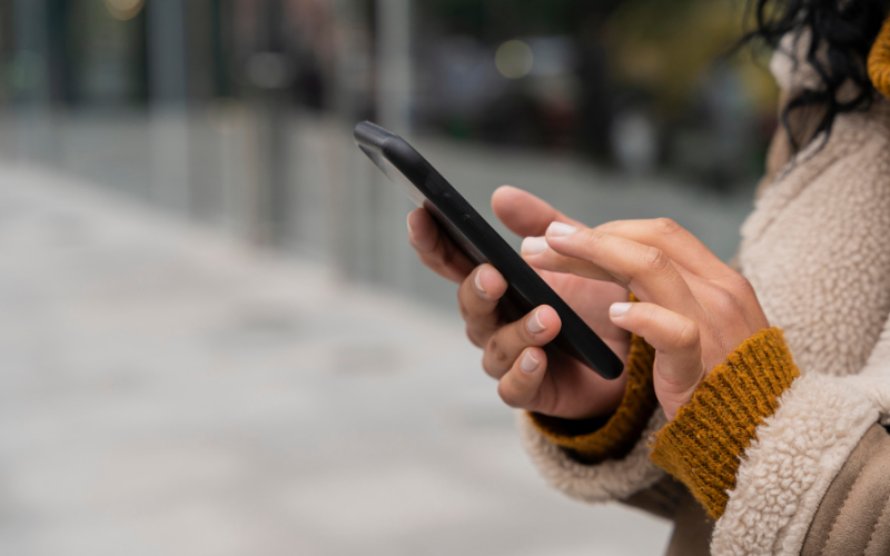 Nahaufnahme von Händen einer Person, die ein Smartphone hält und den Bildschirm berührt; unscharfer Hintergrund im Freien.