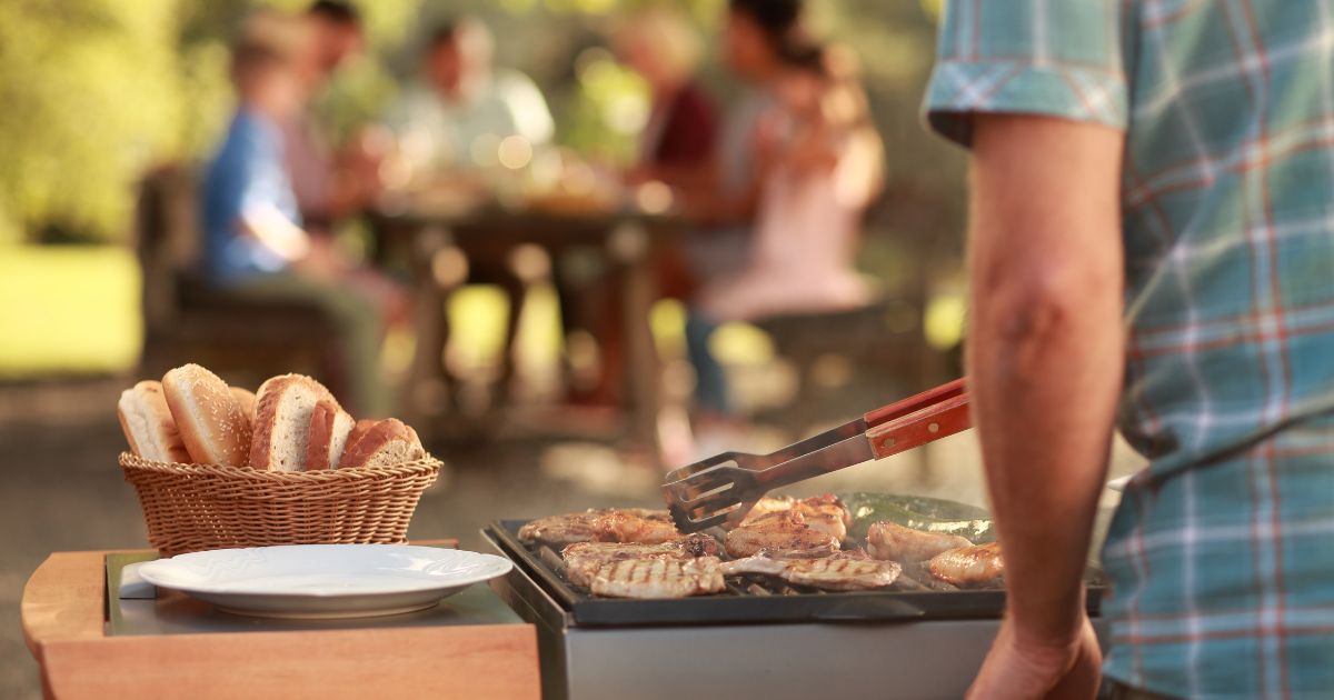 Mann grillt Fleisch, im Hintergrund Familie