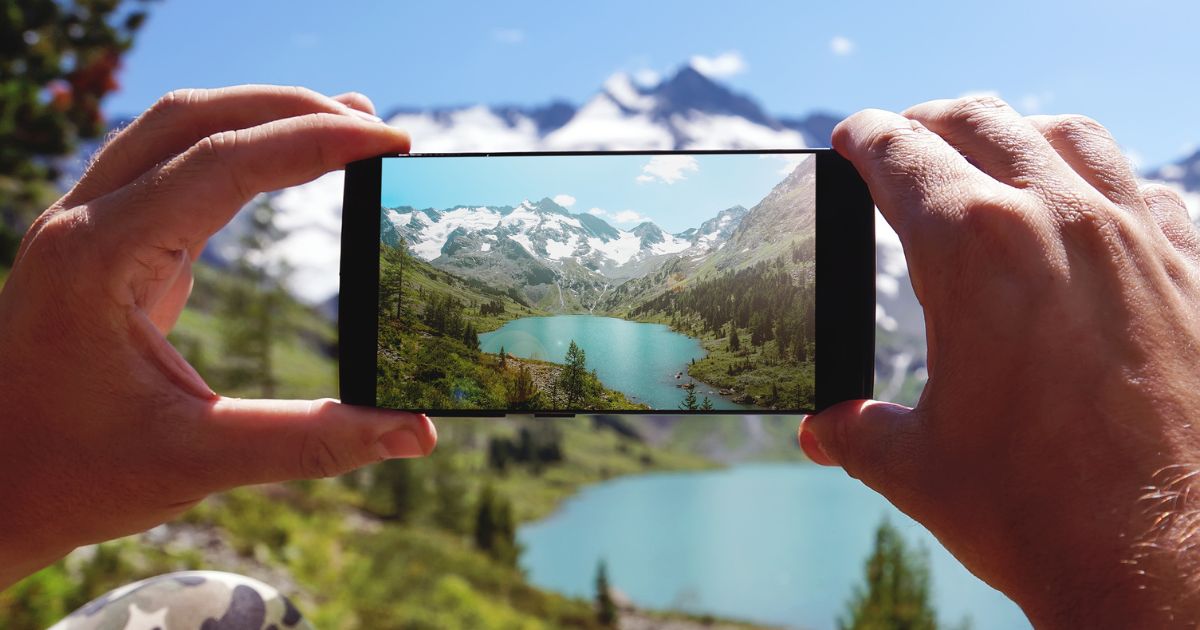 Mensch macht Foto mit Smartphone, im Hintergrund Berge, Wälder und See